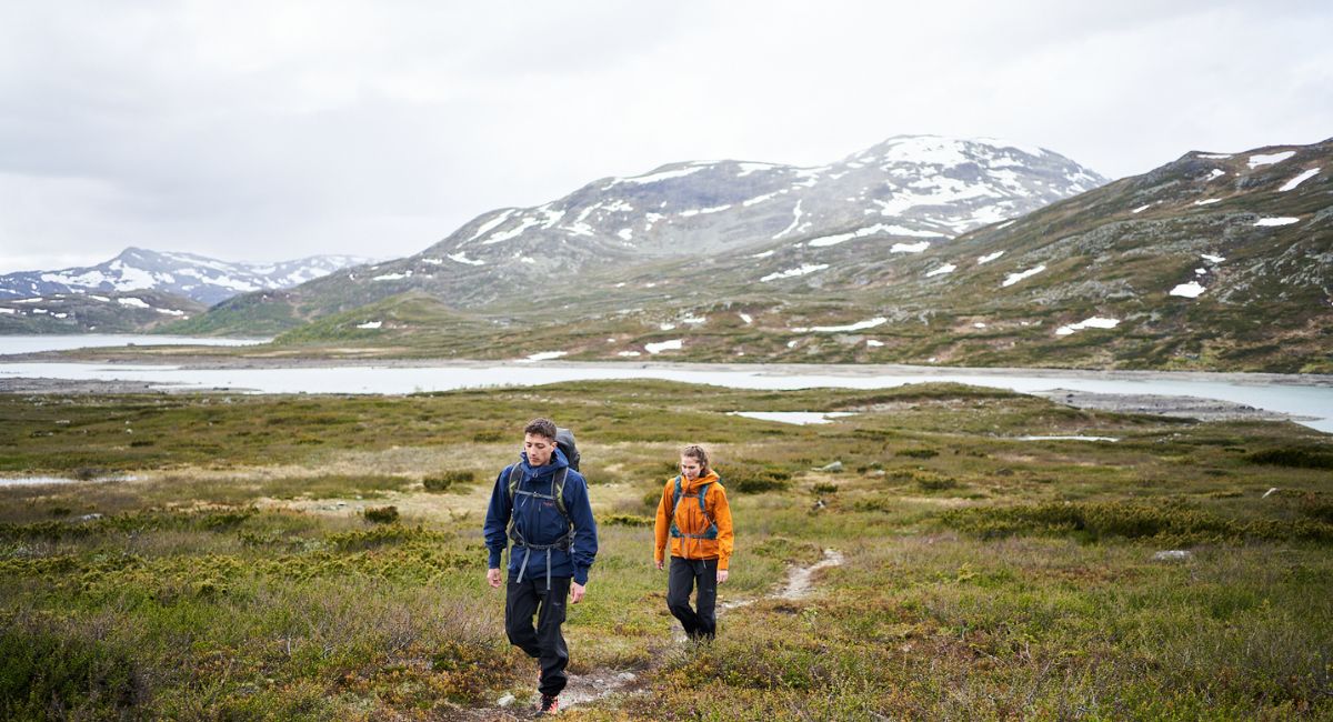 A woman in a bright orange jacket smiles slightly as she zips it up to the top, forest behind in the background